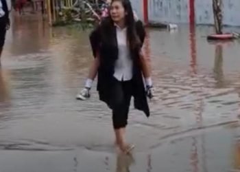 Seorang guru mendongko anak muridnya saat melewati halaman sekolah yang dipenuhi air, Rabu 12 Juli 2023. (Foto: Ist./Koranpapua.id)