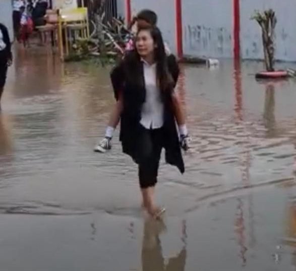 Seorang guru mendongko anak muridnya saat melewati halaman sekolah yang dipenuhi air, Rabu 12 Juli 2023. (Foto: Ist./Koranpapua.id)